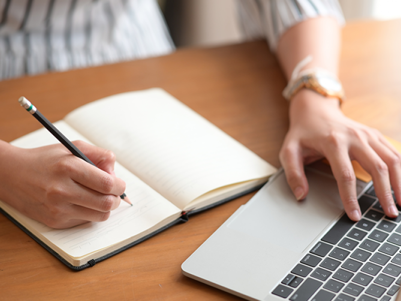 A woman's hands - one hand is writing notes in a notebook, the other is typing on a laptop.
