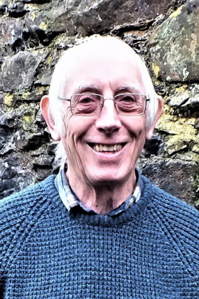 Colin Luker, wearing a knitted jumper and glasses, standing outside in front of a stone wall
