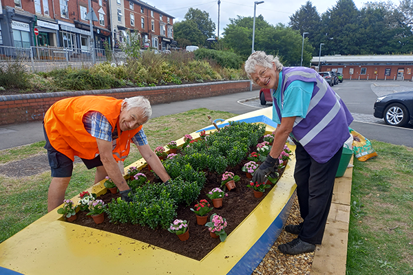 Wyre Forest u3a members Teresa and Roy standing over an old boat that has been refurbished into a planter. Their hands are in the soil; they are gardening.