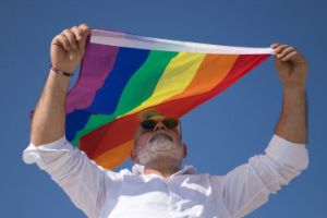 A man with a grey beard wearing a white shirt holding up a pride flag against a blue sky