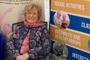 A woman in a pink top and floral jacket sitting down next to a blue and yellow poster